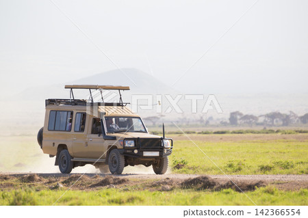 A beige 4x4 safari truck with a raised pop-up roof driving along a dirt road through the open grassy savanna plains of an African national park during a game drive excursion. A beige 4x4 safari truck with a raised pop-up roof driving along a dirt road through the open grassy savanna plains of an African national park during a game drive excursion. 142366554