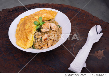 homemade Thai style spicy fried rice with stir-fried chicken and basil cover by a half circle omelet in white plate with spoon in white napkin on wood Serving trays with dark gray background homemade Thai style spicy fried rice with stir-fried chicken and basil cover by a half circle omelet in white plate with spoon in white napkin on wood Serving trays with dark gray background 142366643