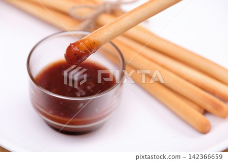 a breadstick dipped with ketchup dipping sauce in transparent cup with pack of breadsticks in background in white dish a breadstick dipped with ketchup dipping sauce in transparent cup with pack of breadsticks in background in white dish 142366659