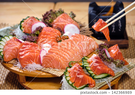 A pair of chopsticks holds a piece of salmon sashimi, against a backdrop of a large sushi set that includes salmon, tuna, and other fish A pair of chopsticks holds a piece of salmon sashimi, against a backdrop of a large sushi set that includes salmon, tuna, and other fish 142366989