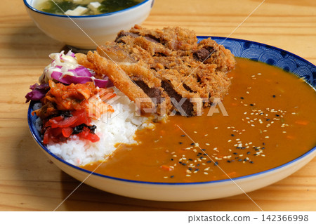 A steaming bowl of Japanese rice with a generous portion of Japanese fried pork and curry sauce, served on a wooden table with a variety of side dishes. A steaming bowl of Japanese rice with a generous portion of Japanese fried pork and curry sauce, served on a wooden table with a variety of side dishes. 142366998