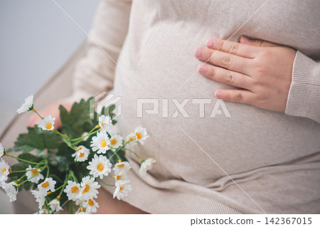 The elderly Asian woman with a pregnant belly sat on the gray sofa, gently cradling her growing baby inside. The elderly Asian woman with a pregnant belly sat on the gray sofa, gently cradling her growing baby inside. 142367015