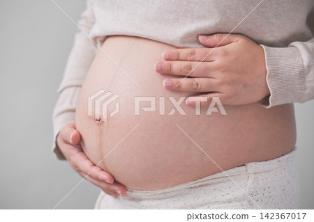 The mature Asian woman stood on the white backdrop, gracefully unveiling her large round belly, cradling it with both of her hands. The mature Asian woman stood on the white backdrop, gracefully unveiling her large round belly, cradling it with both of her hands. 142367017