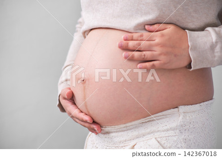 The Asian woman with a pregnant belly stood on the white backdrop, gracefully unveiling her sizeable round belly and cradling it with her hand. The Asian woman with a pregnant belly stood on the white backdrop, gracefully unveiling her sizeable round belly and cradling it with her hand. 142367018