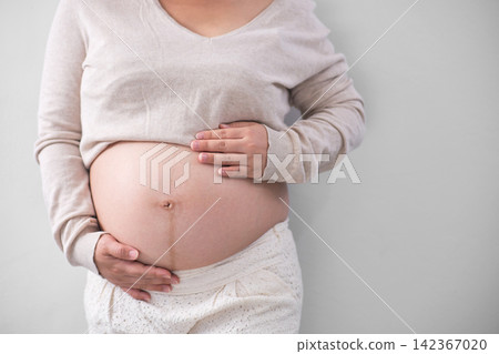 The Asian woman with a pregnant belly stood on the white backdrop, gracefully unveiling her sizeable round belly and cradling it with her hand. The Asian woman with a pregnant belly stood on the white backdrop, gracefully unveiling her sizeable round belly and cradling it with her hand. 142367020