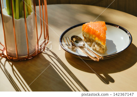 A slice of orange cake, sitting on a dish, is served on a table in a cafe next to a vase of flowers. The soft afternoon sunlight streams in through the window, creating a warm and inviting atmosphere. A slice of orange cake, sitting on a dish, is served on a table in a cafe next to a vase of flowers. The soft afternoon sunlight streams in through the window, creating a warm and inviting atmosphere. 142367147