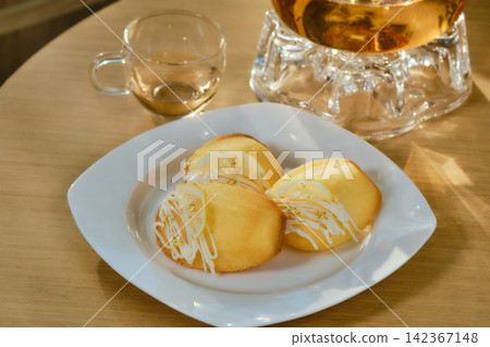 A dish of madeleines with fork and clear glasses jar of herbals tea set on wooden table decorated by flowers blow and a book beside the leaning area with pillow in background A dish of madeleines with fork and clear glasses jar of herbals tea set on wooden table decorated by flowers blow and a book beside the leaning area with pillow in background 142367148