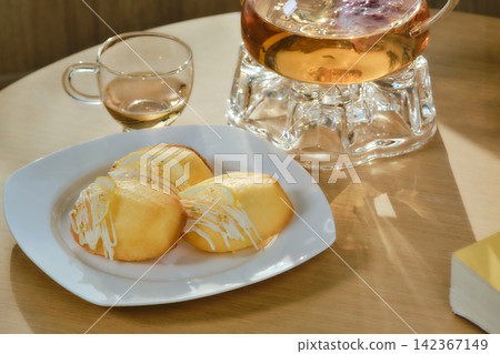 A dish of madeleines with fork and clear glasses jar of herbals tea set on wooden table decorated by flowers blow and a book beside the leaning area with pillow in background A dish of madeleines with fork and clear glasses jar of herbals tea set on wooden table decorated by flowers blow and a book beside the leaning area with pillow in background 142367149