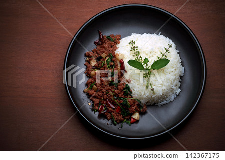 top view shot of home made Thai's style spicy stir fried basil with minced beef served with Thai jasmine rice in black dish put on dark red wooden table with small free copy space on a side top view shot of home made Thai's style spicy stir fried basil with minced beef served with Thai jasmine rice in black dish put on dark red wooden table with small free copy space on a side 142367175
