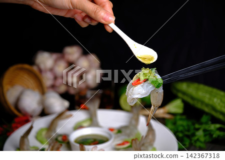 chopsticks tong a fresh raw prawn chili and basil with another hand pouring spicy seafood sauce with a dish and seasonal in background chopsticks tong a fresh raw prawn chili and basil with another hand pouring spicy seafood sauce with a dish and seasonal in background 142367318
