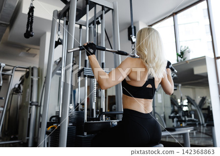 Athletic woman exercising back muscles using lat machine in gym Athletic woman exercising back muscles using lat machine in gym 142368863
