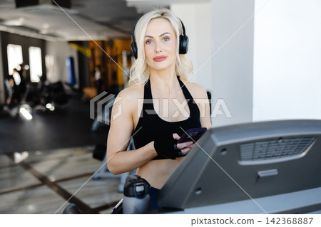 Athletic woman using smartphone and wearing headphones on treadmill in gym Athletic woman using smartphone and wearing headphones on treadmill in gym 142368887