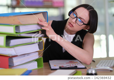 Concerned businesswoman steadies tall binder stack at office desk Concerned businesswoman steadies tall binder stack at office desk 142369432