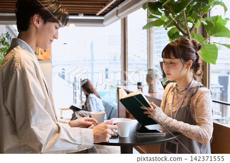 一位年輕女子正在咖啡館裡看書,一名男子坐在她對面。 一位年輕女子正在咖啡館裡看書,一名男子坐在她對面。 142371555