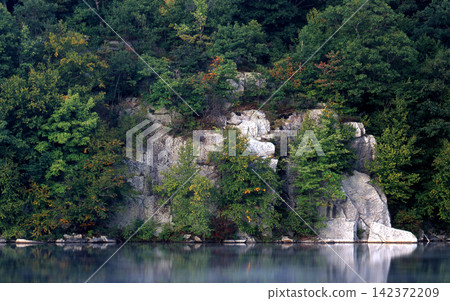 Forested Rocky Lake Shore with Calm Water Reflection Forested Rocky Lake Shore with Calm Water Reflection 142372209
