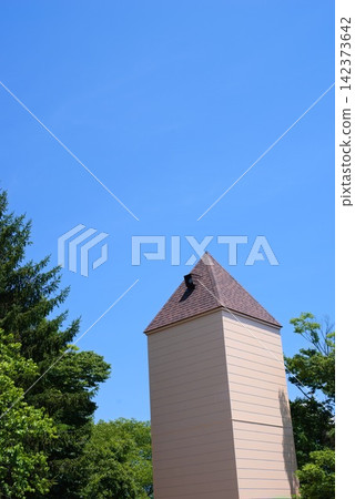 Architectural landscape image featuring a tower-like building and green trees against a wide blue sky. Architectural landscape image featuring a tower-like building and green trees against a wide blue sky. 142373642