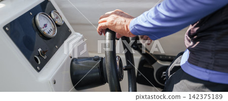 Person holding steering wheel while navigating boat on water, operating maritime vessel during marine journey, captain driving transport vehicle with gauges visible on dashboard panel console Person holding steering wheel while navigating boat on water, operating maritime vessel during marine journey, captain driving transport vehicle with gauges visible on dashboard panel console 142375189