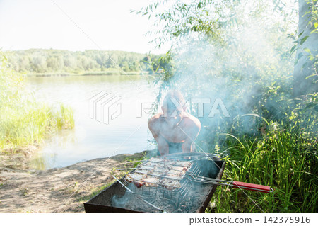 A young teenage boy stands behind a grill with his silhouette partially hidden by thick white smoke rising from juicy meat cooking on a wire rack. A young teenage boy stands behind a grill with his silhouette partially hidden by thick white smoke rising from juicy meat cooking on a wire rack. 142375916
