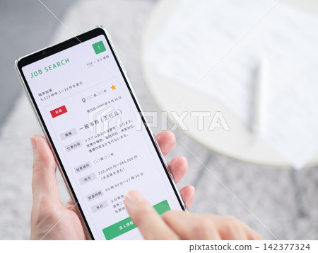A photo of a woman's hands searching for an office job on a smartphone job search website (job search/career change). The smartphone screen content is original. A photo of a woman's hands searching for an office job on a smartphone job search website (job search/career change). The smartphone screen content is original. 142377324
