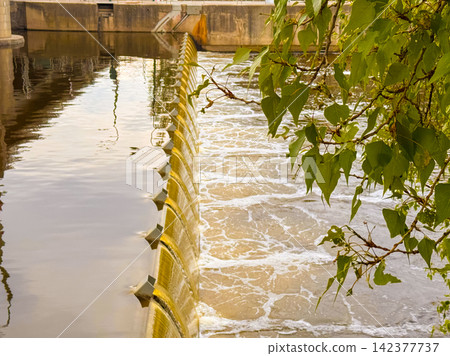 Water flows over a dam near a city on a sunny day Water flows over a dam near a city on a sunny day 142377737