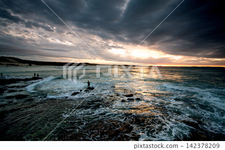 Stormy Ocean Sunset Over Rocky Coast with Fishermen Silhouettes Stormy Ocean Sunset Over Rocky Coast with Fishermen Silhouettes 142378209