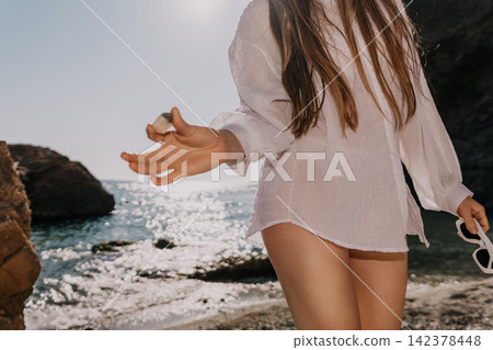 Woman beach fashion Young woman wearing white linen shirt holds a stone on a sunny coast with ocean waves Woman beach fashion Young woman wearing white linen shirt holds a stone on a sunny coast with ocean waves 142378448