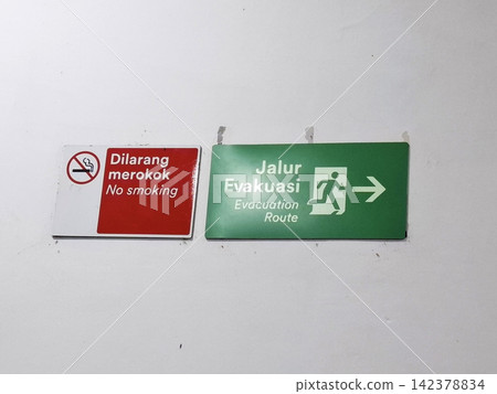 Wall mounted safety signs including red no smoking icon and green evacuation route arrow for emergency building exit. Wall mounted safety signs including red no smoking icon and green evacuation route arrow for emergency building exit. 142378834
