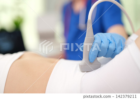 performing ultrasound on patient's abdomen in medical setting with professional medical gloves and blue uniform close-up perspective focusing on ultrasound probe. performing ultrasound on patient's abdomen in medical setting with professional medical gloves and blue uniform close-up perspective focusing on ultrasound probe. 142379211