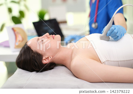 Woman undergoing ultrasound examination of breast, in medical office, with ultrasound machine, medical equipment, pillow, from a side view perspective, with blue, white accents. Woman undergoing ultrasound examination of breast, in medical office, with ultrasound machine, medical equipment, pillow, from a side view perspective, with blue, white accents. 142379222