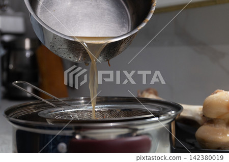 Pouring liquid from metal pot through mesh strainer into receiving pot in kitchen Pouring liquid from metal pot through mesh strainer into receiving pot in kitchen 142380019