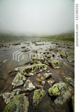 Mossy boulders in shallow clear water of foggy mountain lake Mossy boulders in shallow clear water of foggy mountain lake 142380102