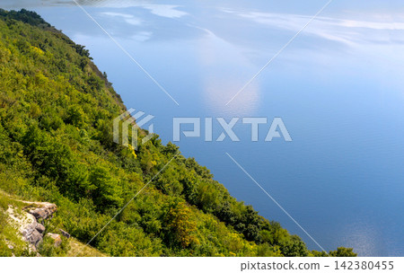Scenic high-angle view of a lush green forested slope meeting a vast blue water body with sun reflection. Nature, landscape, travel, and summer outdoor concept Scenic high-angle view of a lush green forested slope meeting a vast blue water body with sun reflection. Nature, landscape, travel, and summer outdoor concept 142380455