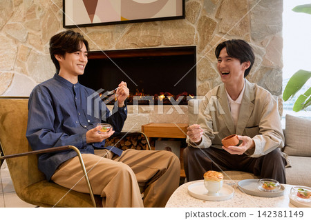 兩個面帶笑容的男人在有壁爐的休息室裡享用甜點。 兩個面帶笑容的男人在有壁爐的休息室裡享用甜點。 142381149