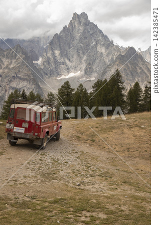 Red off-road vehicle driving mountain trail with peaks and clouds Red off-road vehicle driving mountain trail with peaks and clouds 142385471
