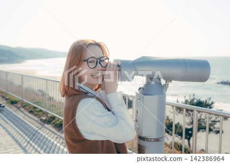 A young woman gazes at the sea through a telescope at an observation deck. A young woman gazes at the sea through a telescope at an observation deck. 142386964