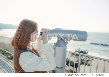 A young woman gazes at the sea through a telescope at an observation deck. A young woman gazes at the sea through a telescope at an observation deck. 142386965
