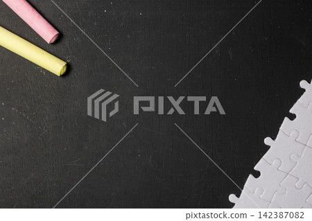 Top view flat lay composition of pink and yellow chalk sticks with white jigsaw puzzle pieces on a dark chalkboard background with copy space. High Quality Photo Top view flat lay composition of pink and yellow chalk sticks with white jigsaw puzzle pieces on a dark chalkboard background with copy space. High Quality Photo 142387082