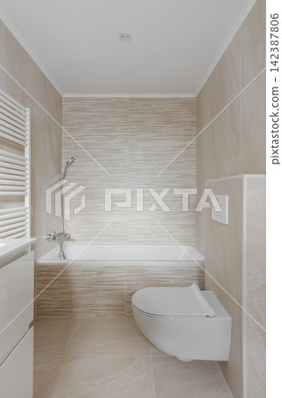 Minimalist modern bathroom interior with bathtub, wall mounted shower, beige textured tile, wall hung toilet, bright neutral finishes, clean residential layout, and refined spa inspired comfort Minimalist modern bathroom interior with bathtub, wall mounted shower, beige textured tile, wall hung toilet, bright neutral finishes, clean residential layout, and refined spa inspired comfort 142387806