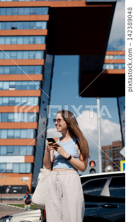 young woman casually texting on phone in urban outdoor scene with modern buildings and blue sky, wearing sunglasses, necklace, and watch, holding white bag, with a relaxed and casual mood. young woman casually texting on phone in urban outdoor scene with modern buildings and blue sky, wearing sunglasses, necklace, and watch, holding white bag, with a relaxed and casual mood. 142389048
