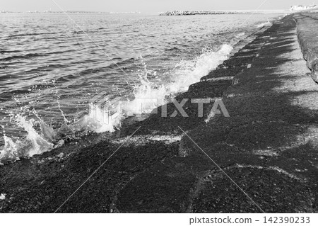 A striking black and white photograph featuring a dynamic coastal scene with a concrete breakwater extending diagonally towards the horizon A striking black and white photograph featuring a dynamic coastal scene with a concrete breakwater extending diagonally towards the horizon 142390233