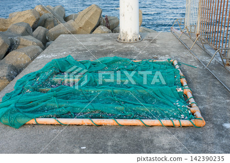 A detailed close-up view of a large rectangular green fishing net on a concrete dock surrounded by ocean waves and rocky breakwater A detailed close-up view of a large rectangular green fishing net on a concrete dock surrounded by ocean waves and rocky breakwater 142390235