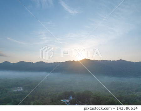 Sunrise emerging behind a forested mountain ridge above misty tropical countryside, with golden morning light illuminating low fog and lush woodland for a peaceful nature. Travel and wellness themes. Sunrise emerging behind a forested mountain ridge above misty tropical countryside, with golden morning light illuminating low fog and lush woodland for a peaceful nature. Travel and wellness themes. 142390614