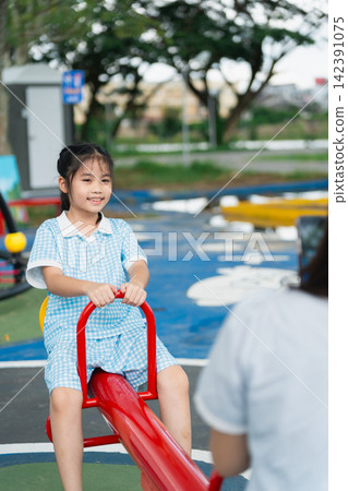 Happy young girl wearing blue checkered dress playing on bright red seesaw at outdoor playground with greenery and urban background during daytime Happy young girl wearing blue checkered dress playing on bright red seesaw at outdoor playground with greenery and urban background during daytime 142391075