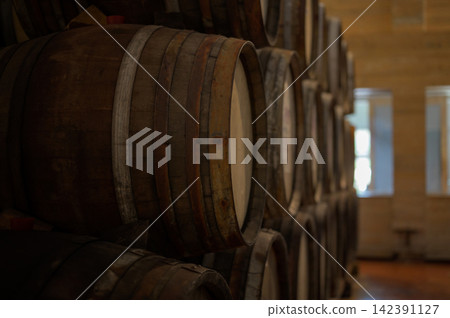 Vintage wooden barrels stacked in rows inside a traditional wine cellar or distillery. Concept of aging process, craftsmanship, alcohol production, and rustic background with warm natural lighting. Vintage wooden barrels stacked in rows inside a traditional wine cellar or distillery. Concept of aging process, craftsmanship, alcohol production, and rustic background with warm natural lighting. 142391127