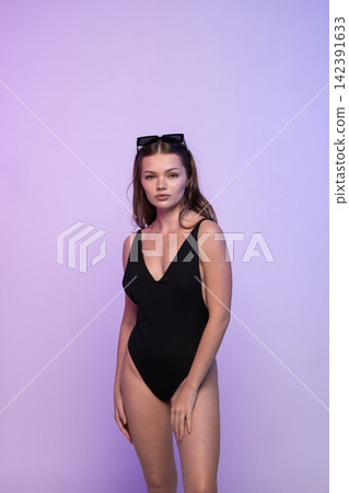 Young woman confidently posing in black swimsuit with sunglasses against purple gradient background Young woman confidently posing in black swimsuit with sunglasses against purple gradient background 142391633