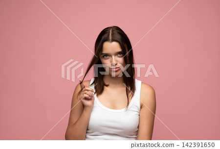Playful brunette woman twirling hair with a flirty expression against pastel pink background in casual white tank top Playful brunette woman twirling hair with a flirty expression against pastel pink background in casual white tank top 142391650