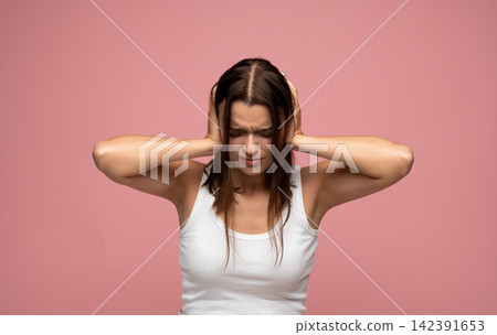 Young woman suffering from headache, covering ears with hands, feeling pain and frustration against a pink background Young woman suffering from headache, covering ears with hands, feeling pain and frustration against a pink background 142391653