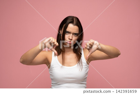 Young woman showing thumbs down gesture expressing rejection and disapproval against a pink background Young woman showing thumbs down gesture expressing rejection and disapproval against a pink background 142391659