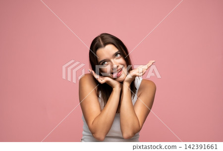 Young woman making a playful silly face sticking out tongue and looking up against pink background Young woman making a playful silly face sticking out tongue and looking up against pink background 142391661