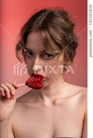 Young woman with red lips seductively holding a heart-shaped lollipop, celebrating love and romance on a pink background 142391816